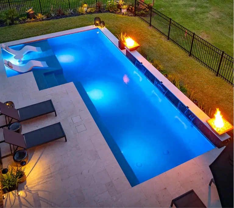 Wide-angle nighttime view of a Legacy Pools luxury remodel featuring glowing blue LED lighting, fire features, and submerged lounge chairs.