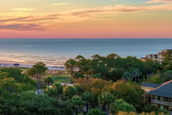 View of a beach with calm ocean waves, sandy shore, trees, and several buildings at sunset with colorful sky.