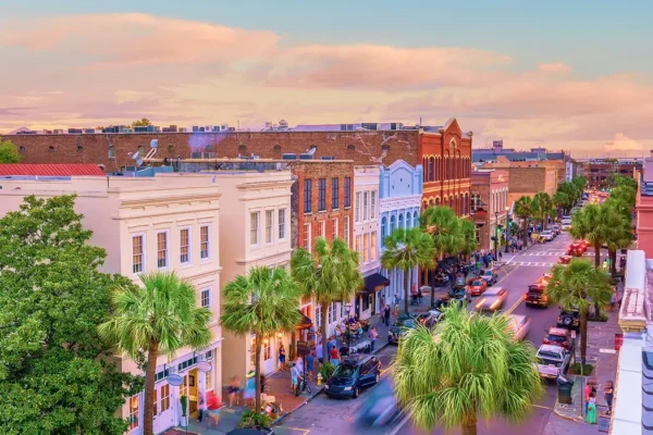 A colorful row of historic buildings lines a busy street with palm trees and cars, under a pastel sunset sky.