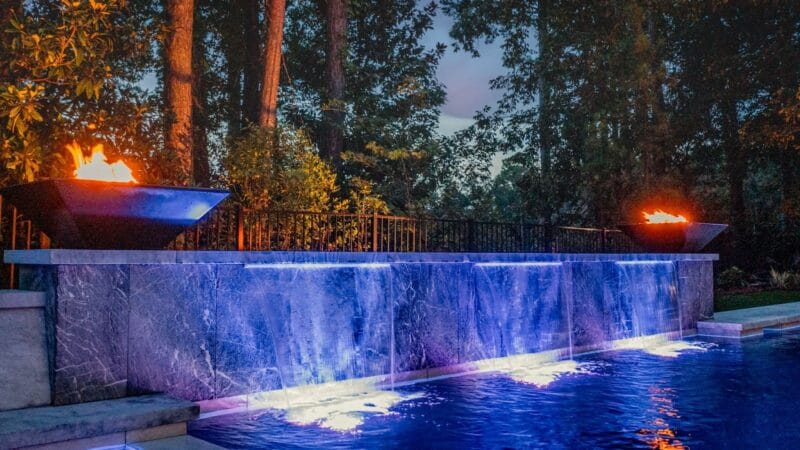 A lit-up pool at night with fire bowls and waterfalls