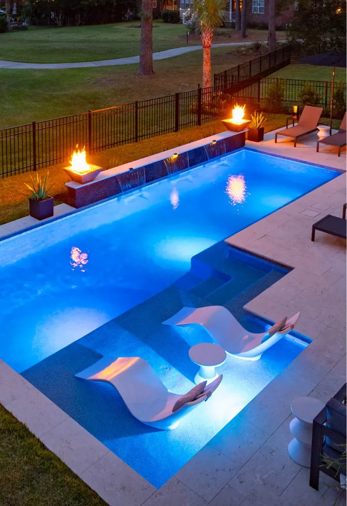 Luxurious outdoor pool at night, glowing blue with two submerged loungers. Fire bowls and deck chairs create a serene, elegant atmosphere.