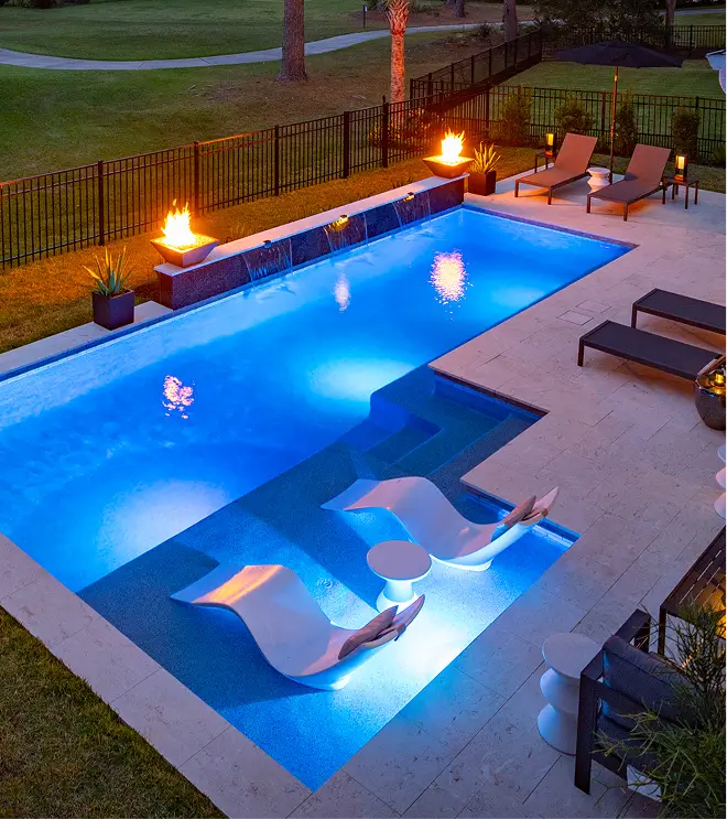 Resort-style pool at dusk with fire bowls and illuminated in-pool lounge chairs.