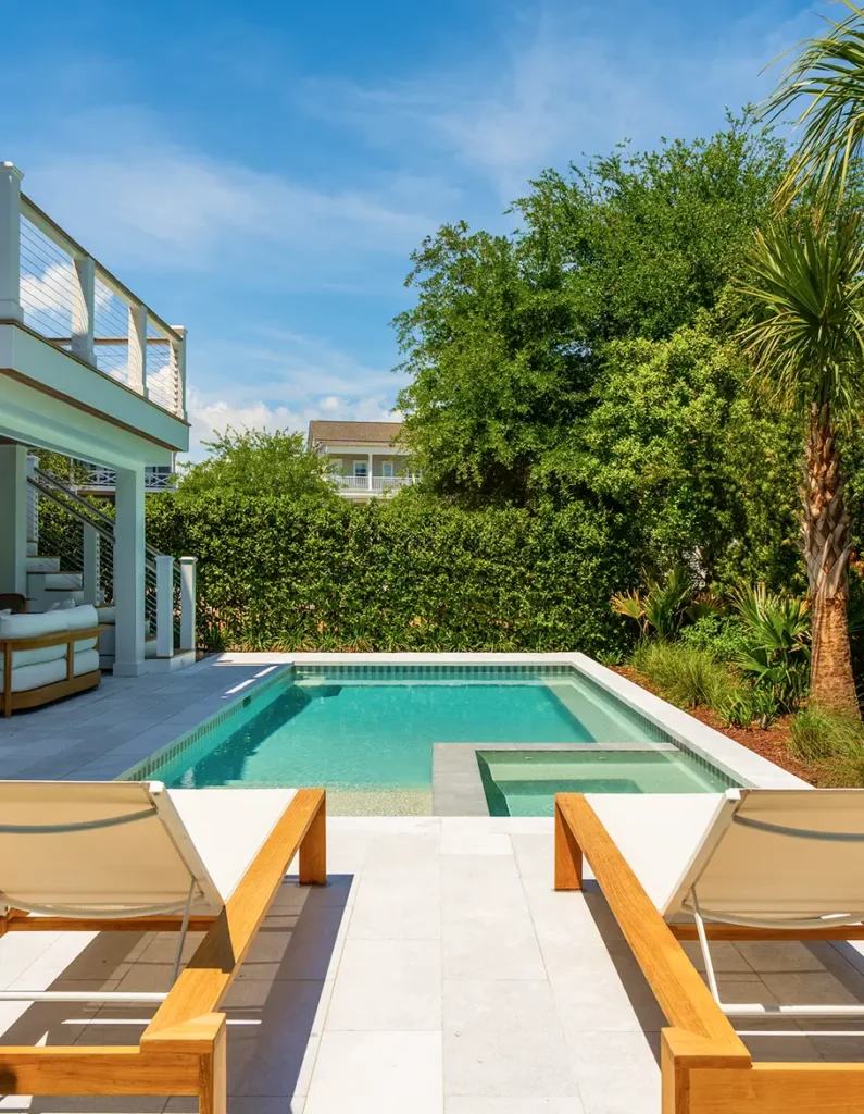 A sunlit backyard with a small, clear pool surrounded by lush greenery. Two wooden lounge chairs face the pool, offering a tranquil, relaxing vibe.