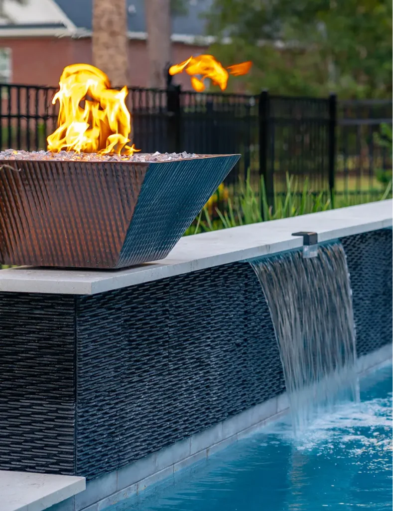 A modern outdoor fire pit with flames sits beside a sleek water feature cascading into a blue pool, creating a serene and inviting atmosphere.